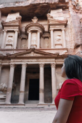Fototapeta na wymiar Young European tourist looking at the Treasury of Petra, Jordan. Brunette girl in red dress enjoying the view of the façade and the ruins of the ancient city carved in stone.