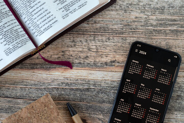 Digital calendar 2024 on smartphone and open holy book on wooden background. Top view. Biblical...
