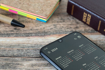 Digital calendar 2024 on smartphone screen with closed holy bible book and notebook on wooden...