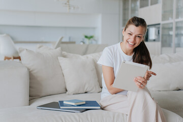  cheerful woman in casual attire enjoys using a tablet on a comfortable sofa in a bright, airy...