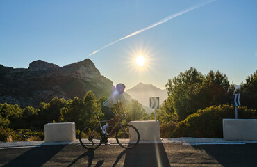 A man is riding a gravel bicycle on the road in the hills with a mountain view at sunset. Athlete...