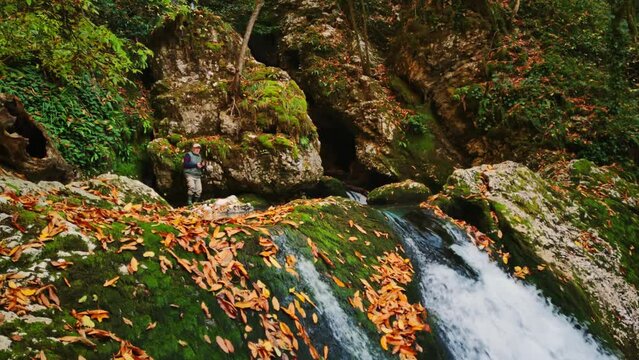 Man is standing on the rock and filming waterfall flowing from it on smartphone in his hands filmed in slow motion at autumn day. Yellow foliage is flying in air and male tourist enjoying landscape