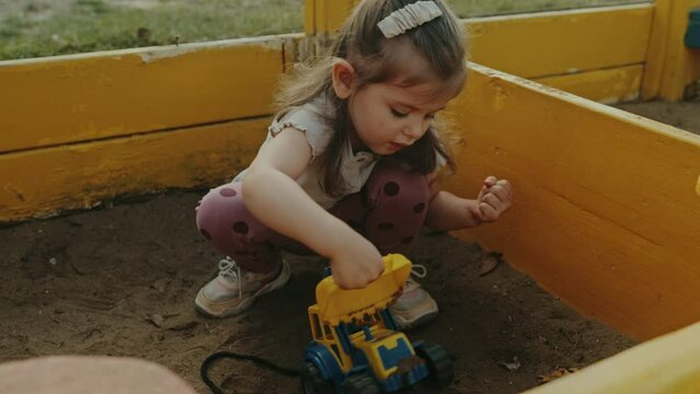 Little happy girl playing with yellow truck toy in a sandbox in the yard on a summer day. Kid having fun.