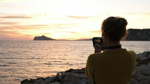 A tourist woman taking photos and videos of the amazing colorful sunset sky on the Mediterranean Sea. Hipster girl taking a summertime break on the coastline, capturing footage for social media. Spain