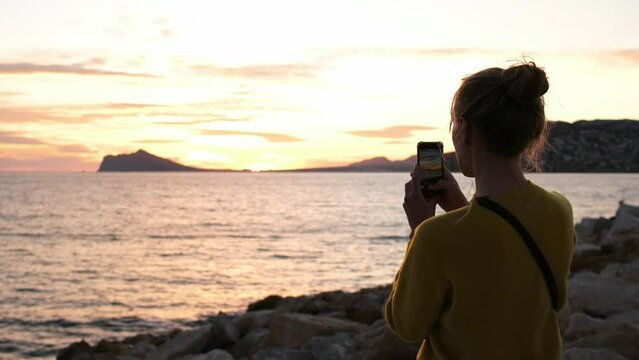 A tourist woman taking photos and videos of the amazing colorful sunset sky on the Mediterranean Sea. Hipster girl taking a summertime break on the coastline, capturing footage for social media. Spain