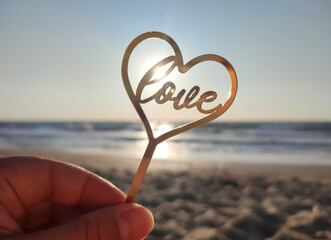Person holding in fingers hand stick in shape golden heart and word Love on background sea and sea...
