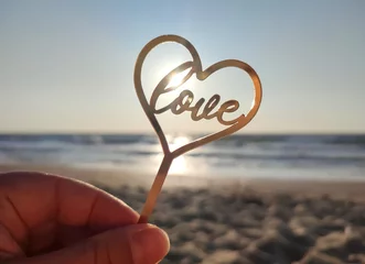 Fotobehang Person holding in fingers hand stick in shape golden heart and word Love on background sea and sea waves in seashore on sunny summer day close-up. Concept love romance amour St Valentines Day © mari1408