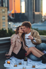 Fototapeta premium Romantic beautiful surprise on Saint Valentine's Day for a girlfriend. Loving young couple having dinner on a rooftop on sunset evening with candles, wine. Skyscrapers on background, urban cityscape