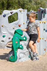 Little cute boy standing in playground by modern gray plastic climbing wall and looking at green...