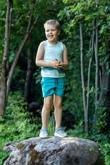 Little boy standing on a big boulder in the summertime. He smile. Beautiful green foliage background.