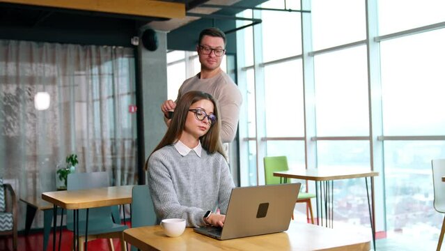 Long-haired female working at her laptop in the office. Man stirring sugar in the cup comes up behind peeping at colleagues computer.