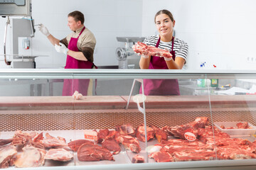 Positive young girl, professional butcher shop saleswoman, arranging meat products in refrigerated...
