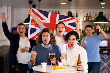 Happy young adult fans waving British flag while drinking beer and watching match together in...