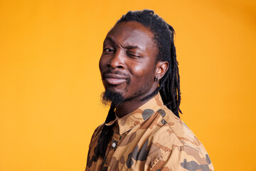 Smiling playful african american man winking at camera, fooling around over yellow background. Portrait of positive young adult having fun doing eye wink in studio, enjoying free time. - obrazy, fototapety, plakaty