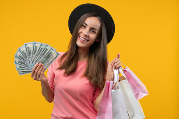 Happy confident smiling young woman posing with shopping bags and ward of dollar banknotes in hands, isolated over yellow background