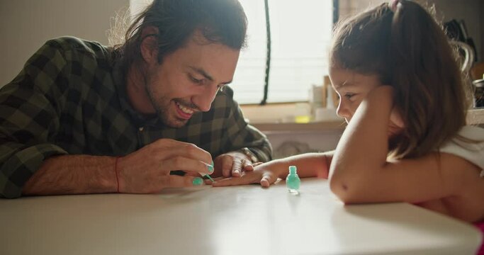A brunette man in a green checkered shirt gives his little daughter a brunette girl in a pink dress a manicure using green polish in the kitchen on the table