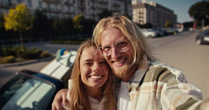 Portrait of a happy couple, a blond guy in glasses with a beard takes a selfie with his blonde girlfriend who smiles widely in a White hoodie. They show the V sign and have fun in the city