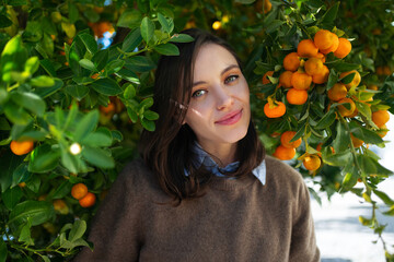 Girl standing under branches of tangerines surrounded citrus fruits. Smiling woman stands under...