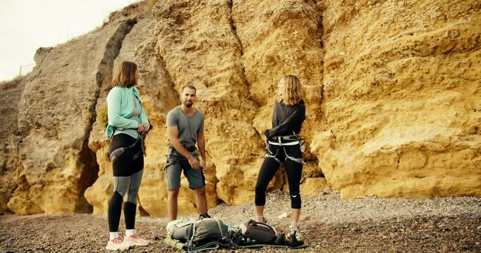 A blonde girl in a blue jacket, a blonde girl with curly hair in black clothes and a brunette guy in a gray T-shirt put on a belay before they start rock climbing on a rocky shore near the yellow