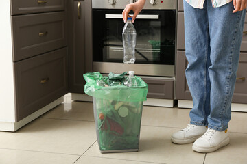 Woman putting plastic bottle into trash bin in kitchen