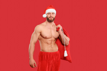 Muscular young man in Santa hat holding bag with presents on red background