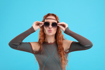 Stylish young hippie woman in sunglasses sending air kiss on light blue background
