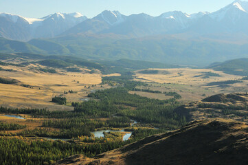 Fototapeta premium The winding bed of a beautiful river with wooded banks flows through the autumn steppe at the foot of a mountain range with snow-capped peaks in the rays of the setting sun.
