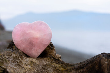 A peaceful image of a large rose quartz crystal heart on a rough piece of brown driftwood and ocean background. 