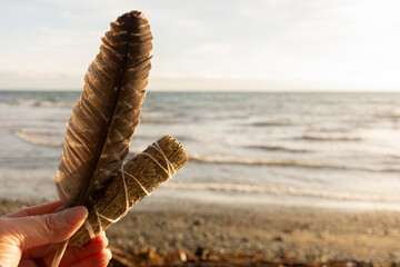 A tranquil image of a hand holding a white sage smudge stick with sacred feather with the Pacific...