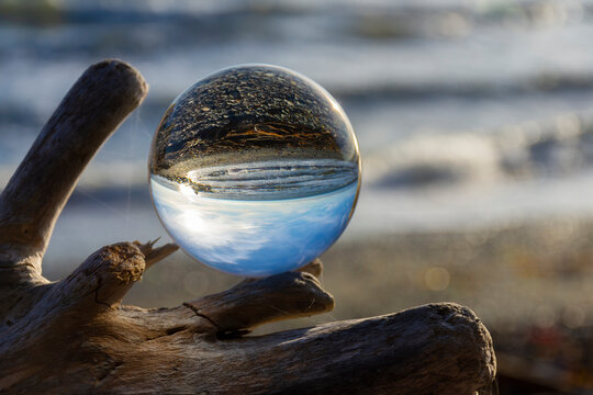 A creative image of a glass lens ball resting on a driftwood log and reflecting an image of the Pacific Ocean shoreline. 