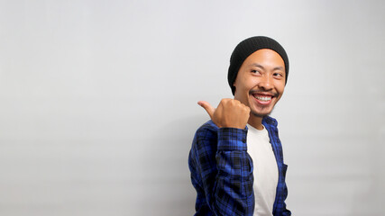 An excited young Asian man, dressed in casual clothes and wearing a beanie hat, is pointing with...