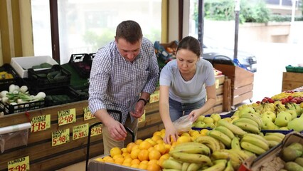 Spouses browsed vibrant display of oranges at supermarket, carefully selecting ripest ones for their homemade pie. Husband and wife came shopping at grocery store and put ripe orange in plastic bag