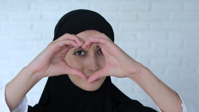 Muslim woman in black hijab depicted heart made of hands. Close-up shot: Muslim woman in black hijab smiles and shows heart from hands. Love and friendship concept, women hands in form of heart