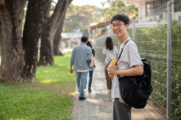 Positive Asian male student with backpack is walking to school, walking on a footpath on his campus.