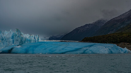 The famous Perito Moreno Glacier. A wall of cracked blue ice rises above the lake. A large breakaway iceberg floats in the water. Coastal mountains in clouds and fog. El Calafate. Argentina.