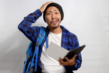 A funny young Asian student, dressed in a beanie hat and casual shirt and carrying a backpack,...
