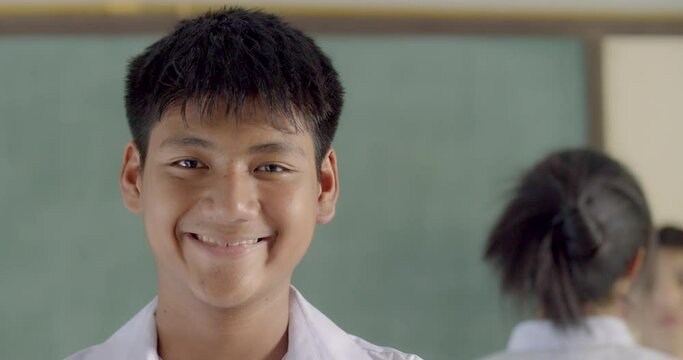A happy smiling male Asian high school student in white uniform standing in classroom.