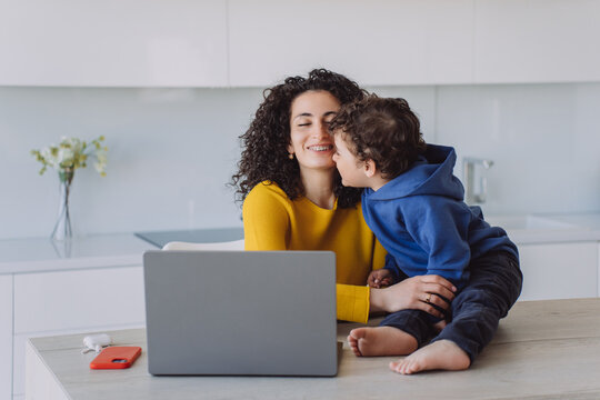 A mother and son share a joyful moment in a bright kitchen, with the mom working on a laptop and the boy expressing love kisses mother, cuddle.