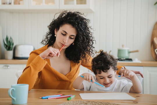 A caring mother in an orange blouse thoughtfully assists her young son with drawing, sharing a creative moment together at their cozy kitchen table.