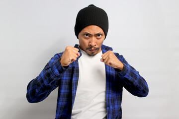 Young Asian man, wearing a beanie hat and casual outfit, is displaying aggression with a defensive gesture, clenched fists, an angry expression, and a readiness to fight, indicating potential anger