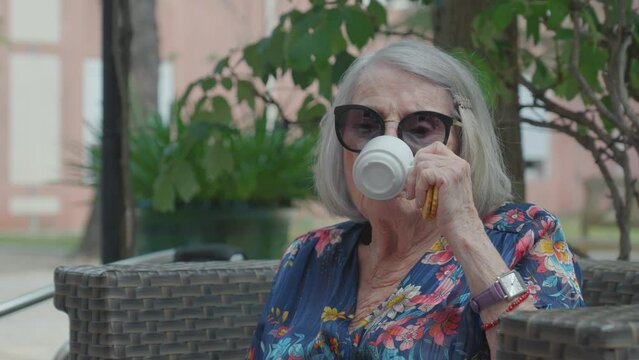 Lovely older woman with sunglasses sips her tea with a cookie in her hand on a sunny day