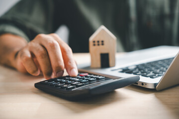 Pressing calculators hand eyes home refinance. Wooden house model, buy or rent note on desk. Saving for property purchase concept efficient mortgage payment. Tax credit analysis for financial strategy - obrazy, fototapety, plakaty