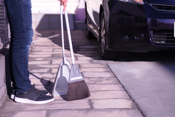 ほうきで外構の掃除をする写真。背景に自動車と玄関ポーチ。家事、家の手入れ、清潔感、ゴミ掃除、管理をイメージ。