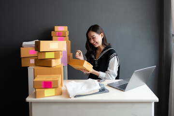 In the office there is Asian female online seller, owner of a startup business, looks at a package, smiling, there is a smartphone, laptop and clothes. Products ready for delivery to customers