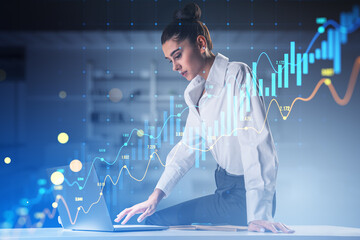Attractive young businesswoman sitting on desk and using laptop with glowing candlestick forex...