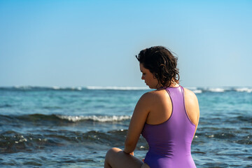 Fototapeta na wymiar Young woman in purple swimsuit sitting if front of ocean.