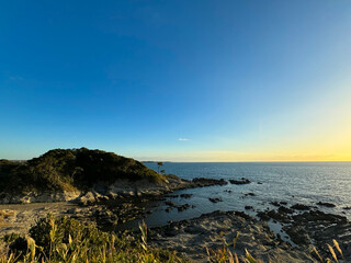 大きな海に波が打ち寄せ、青い大空と太陽と夕日が混ざり合い、高台から見下ろす実写写真