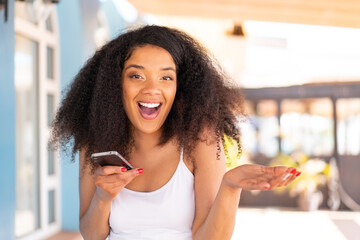 Young African American woman using mobile phone at outdoors with shocked facial expression