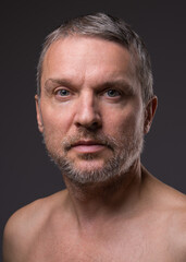 Obraz premium .Slightly smiling face of a middle-aged man with a beard looking at the camera. Close-up portrait of a fifty-year-old man with a naked torso against a dark background.