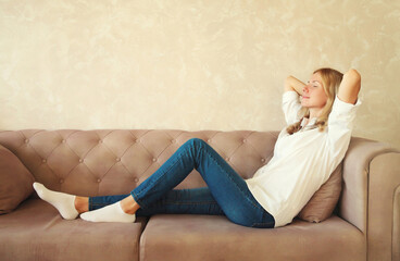 Relaxed caucasian young woman resting with closed eyes and folded hands behind the head while lying on couch in the living room at home